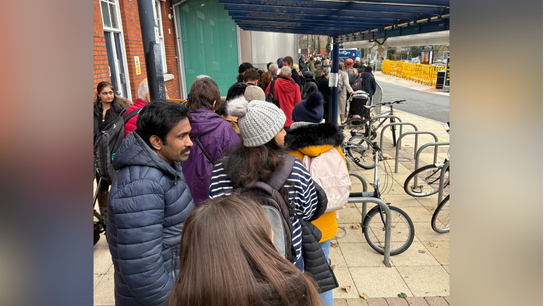 A long queue at Basingstoke Station for rail replacement bus services due to the storm. Adults and children wrapped in warm clothes are lining up around the bus stop. There are a few bikes parked near them.