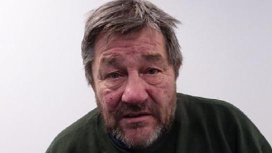 A man of around 60 pictured in a police mugshot. He appears to be hunched, has grey hair and a short beard and is wearing a dark top.