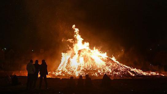 A bonfire which is lit on a dark evening with some people standing around it.