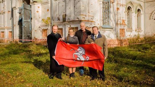 Four people, three men and a woman, standing in front of a white building with bullet holes in it while holding the Kent flag, which is red with a white horse on it