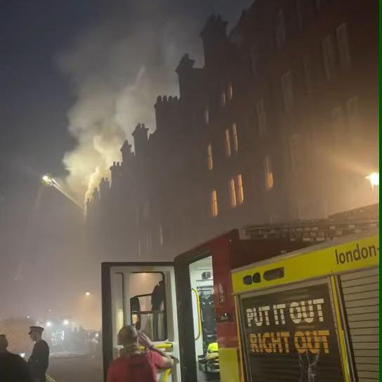 A night time scene of a fire in a multi-story building with thick smoke billowing from the roof. Firefighters are actively responding, with a fire engine in the foreground displaying the slogan "PUT IT OUT RIGHT OUT." A firefighter is seen near the open vehicle, while a high-pressure hose sprays water onto the burning building. Several people, including emergency responders and onlookers, are present.