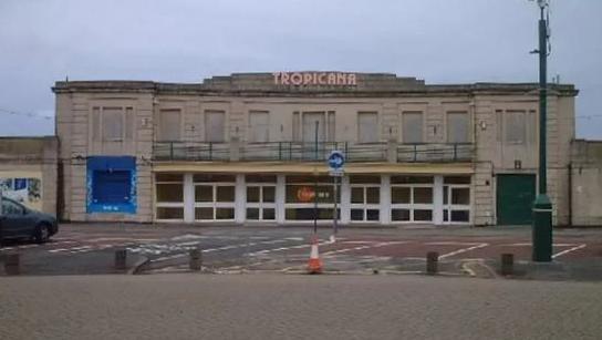 Entrance to cream building, which has Tropicana letters on in rose cold. There are lots of glass doors with white rims. Outside the building is a car park and cone.