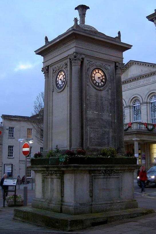 An external view of the Sim's Clock, a four-sided stone tower with a clock face at the top quarter of each side. It is pictured at low light, with a car, a couple of people and a large building behind it.