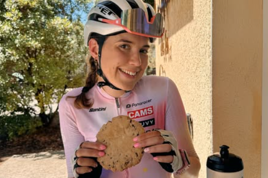 Mrs Hudson wearing pink and white cycling gear, sitting down eating a biscuit. There is a water bottle on the table in front of her.
