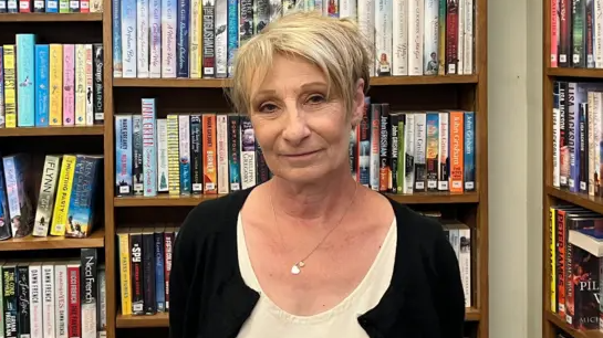 A smiling Jennie Storey looks directly at the camera as she stands in front of a bookshelf full of titles. She is wearing a black cardigan and a white shirt underneath it. She is also wearing a silver necklace with a pendant.