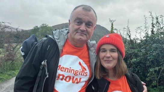 Lloyd's parents, Tony and Louise Sutton in black jackets and oranges t-shirts with the logo of Meningitis Now. They are looking at the camera and Tony is hugging Louise over the shoulder. There is a path and a hill behind them.