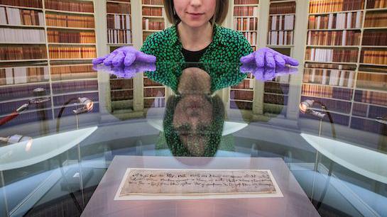 Archivist Jocelyn Grant looks down at the letter in its case. She is wearing top with green and black dots, and purple gloves to handle the letter. Behind are shelves and shelves of bookcases.