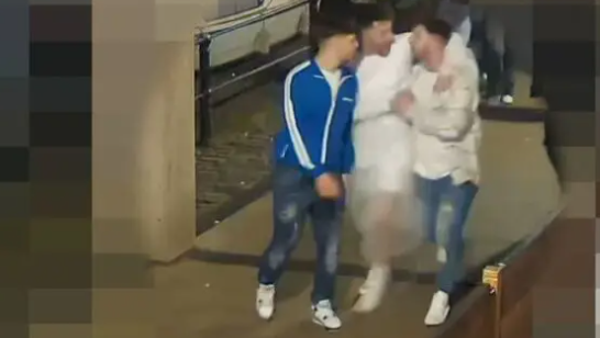 A CCTV image of three men walking under a covered section of Bristol's Harbourside. They all have their arms around each other and are laughing. The man in the middle appears to be wearing a white wedding dress, while the two men either side are in jeans and casual clothes  