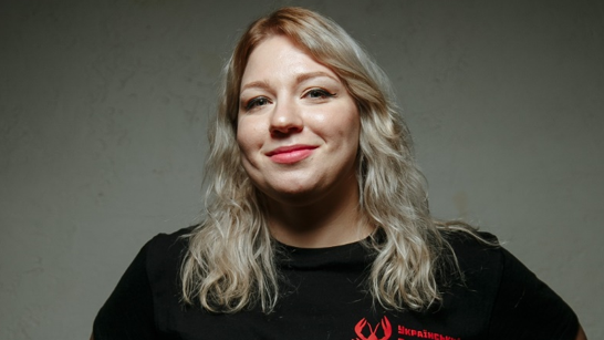 Hanna Kochehura, a blonde lady wearing winged eyeliner, smiles at the camera wearing a black t-shirt. 