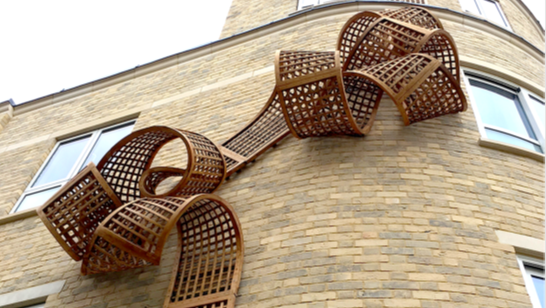 A piece of artwork that looks like a long, thin wooden trellis curled around itself, fixed to the outer wall of a building