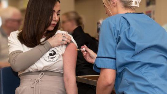 a woman with short dark hair holding the sleeve of her white t-shirt up as a woman, a nurse, wearing blue scrubs inserts a vaccination into her arm