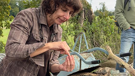 Tara Downs sat in nature, looking at something in her hand. Behind her a man is standing looking on. We cannot see his face. Tara is wearing a brown flannel shirt and sat next to a light blue wheel barrow.