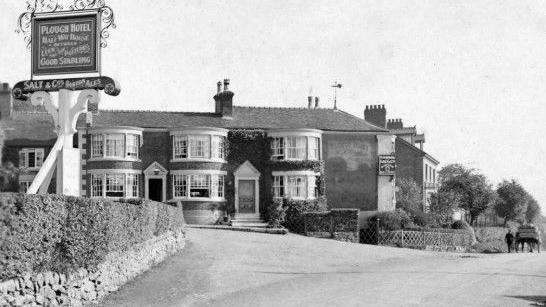 A black and white photograph of The Plough in Endon. There are three bay windows on the ground and first floors of the building. There is a person walking alongside a horse and cart on the right hand side of the image.