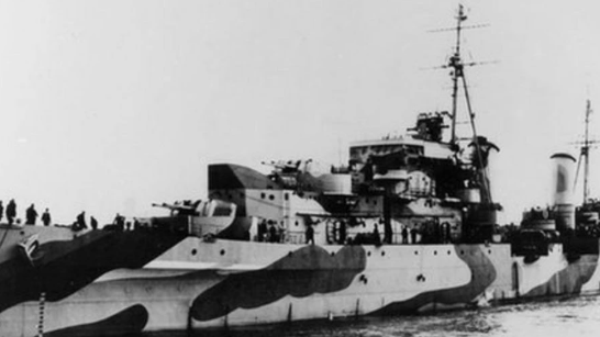 A World War Two warship in black and white. It carries heavy artillery and has a large mast