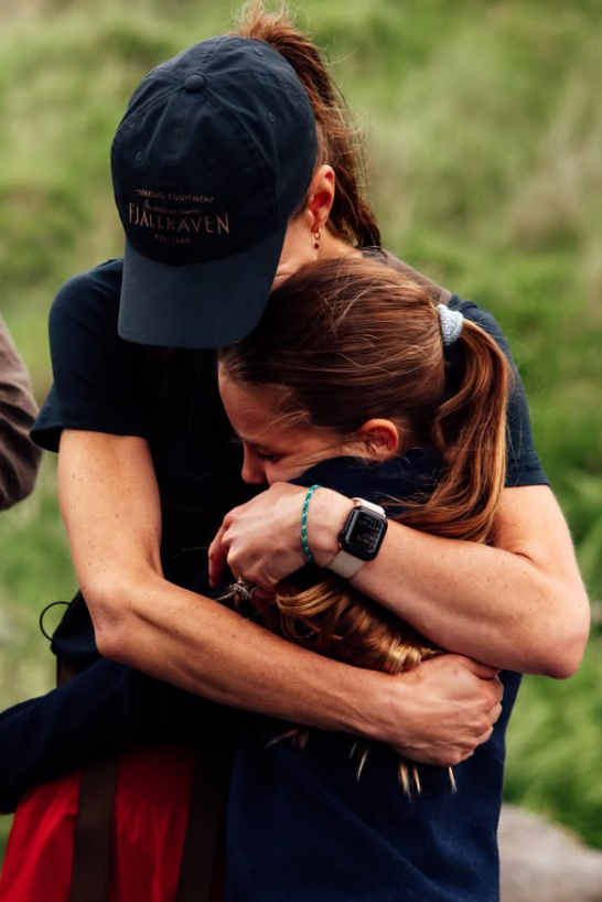 Catherine hugs her daughter Charlotte. Catherine wears a black baseball cap, a black t-shirt and red shorts. Charlotte has long brown hair tied up in a ponytail. Behind them is a green hillside.