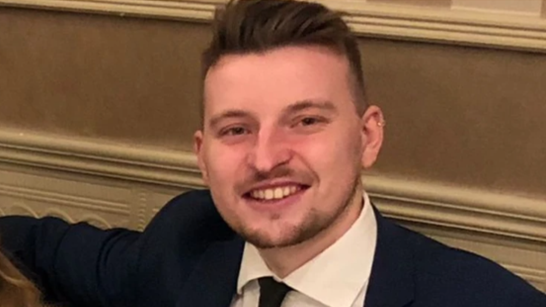 A picture of a man with short, brown, spikey hair, who is wearing a suit and smiling