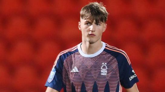 Justin Hanks in action for Nottingham Forest's under-21 side. He is wearing a pink and blue patterned shirt with navy blue short sleeves and three pink stripes running down the shoulders. He is walking and looking into the distance. The background is red showing the empty seats in the stand.
