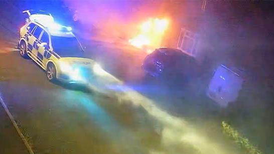 Flames engulf the front of a house with a police car parked outside.