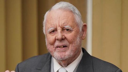 Sir Terry Waite smiles at the camera after receiving a Knighthood at Buckingham Palace in 2023. He has grey hair and a grey beard. He is wearing a dark grey suit with a white shirt and white tie. 