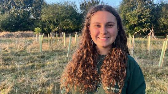 Eden Sedman, a young woman, stands in a grassy meadow with saplings and trees in the background. She has long, curly, auburn hair, wears a green shirt and is smiling into the camera.