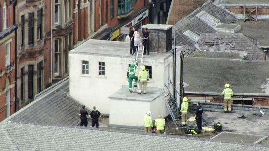 A wide angle shot of a building roof showing fire service personnel, ambulance staff and police officers looking up towards a group of unidentified people standing on a higher section of the roof
