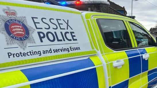 A police van takes up the whole picture. The van is yellow and blue chequered pattern, and has a sign on the back saying Essex Police Protecting and serving Essex. 