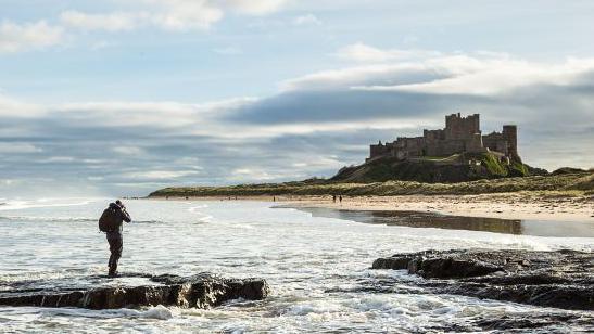 Photographer standing in the surf taking pictures of a cloud-covered Bamburgh Castle  on the Northumberland coast 