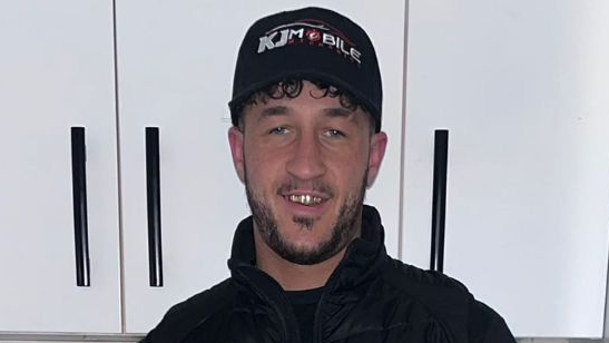 A photograph of Kaine Fletcher, a mixed-race man in his 20s, wearing a black cap which reads 'KJ Mobile' and a black jacket. He is standing in front of white cabinets.