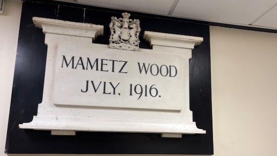 A white marble commemorative plaque with black writing hanging above the entrance to the  Mametz Wood Ward, at Cardiff Royal Infirmary. It shows a crest with a lion and unicorn and reads 'Mametz Wood July 1916'