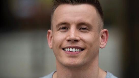 A smiling Lewis Kemp with cropped black hair and wearing a grey jumper