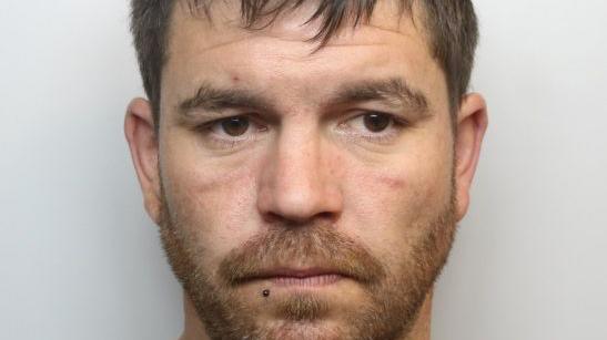 A custody image of Kane Thomas. He has brown hair and ginger beard and is looking off to the right of the image. 