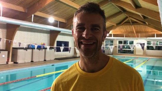 A smiling Steve Harris standing next to the swimming pool. He has short dark hair and is wearing a yellow t-shirt.