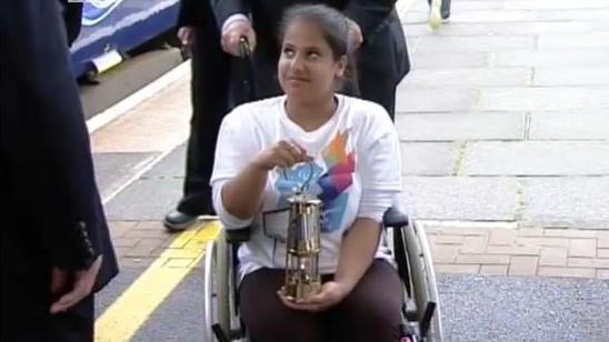 Doaa has her dark hair tied back. She is wearing a white shirt and red trousers. She is seated in a wheelchair and is carrying a lit lamp