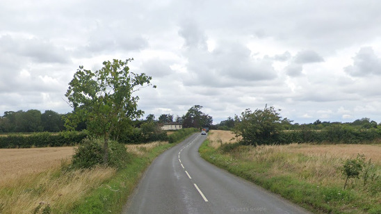 Tivetshall Road in Pulham Market, showing broken white lines on a country road flanked by fields, hedgerows and a tree. A single car can be seen in the distance. 