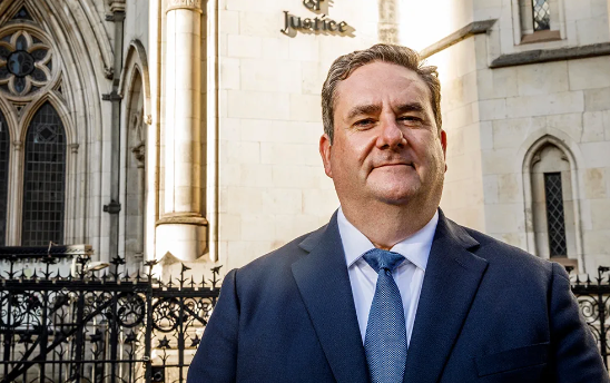 Law Society president Nick Emmerson wearing a blue suit standing outside the Royal Courts of Justice - a pale stone building with wrought iron ornate railings