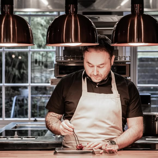 Stuart Ralston works at a bench in the kitchen, with tweezers placing the finishing touches to a food creation. He wears a cream apron. The lighting is moody with three pendant lights above, and windows behind him looking out into a garden area.
