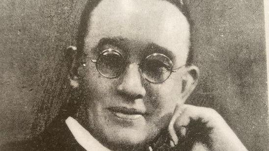 An old black-and-white portrait of a man in a priest's white collar posing with a hand on his chin. He is smiling and wearing round glasses and his dark hair is swept back from his face.