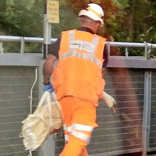 A swan being removed in a bag by a man wearing an orange vest and white hardhat.