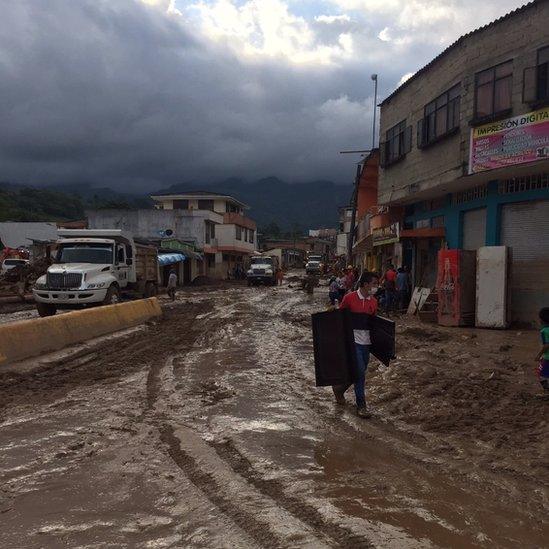 A man carries his possessions through muddied, boarded up street. the tire tracks are thick with mud and the clouds over are gloomy.