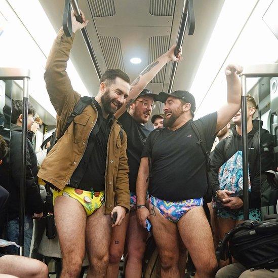 Three men holding a handrail on a Tube train while wearing coconut and neon patterned pants.