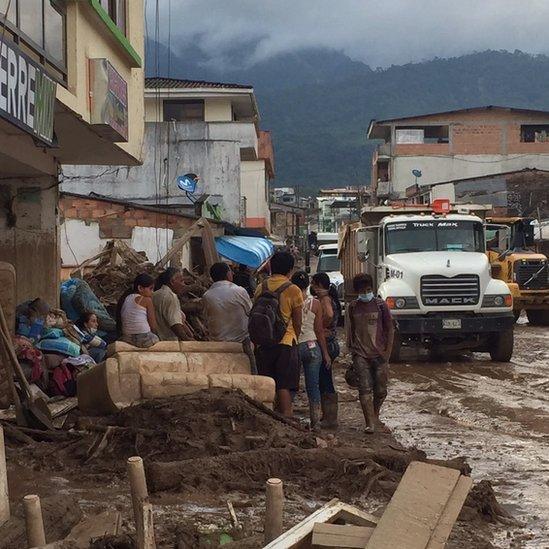 People sit amongst debris and removed possessions. Their legs are drenched with mud and they sit amongst bags and items they have salvaged.