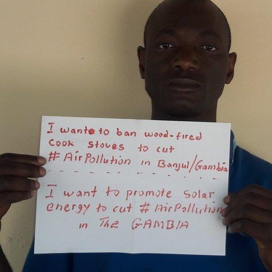 Adboulie Sey holding a sign saying "I want to ban wood fired cook stoves to cut #AirPollution in Banjul, Gambia. I want to promote solar energy to cut #AirPollution in the Gambia."