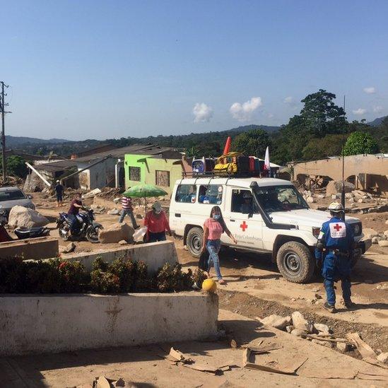 A red cross van is shown with people walking in breathing masks past it through the rubble