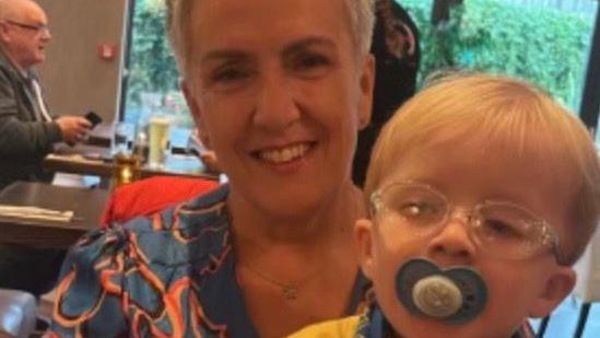 Fiona Scott with her grandson Frankie on her lap. She is smiling at the camera and has grey, cropped hair and a dress on. Frankie is wearing a yellow top, with a blue patterned scarf and he has a dummie in his mouth.