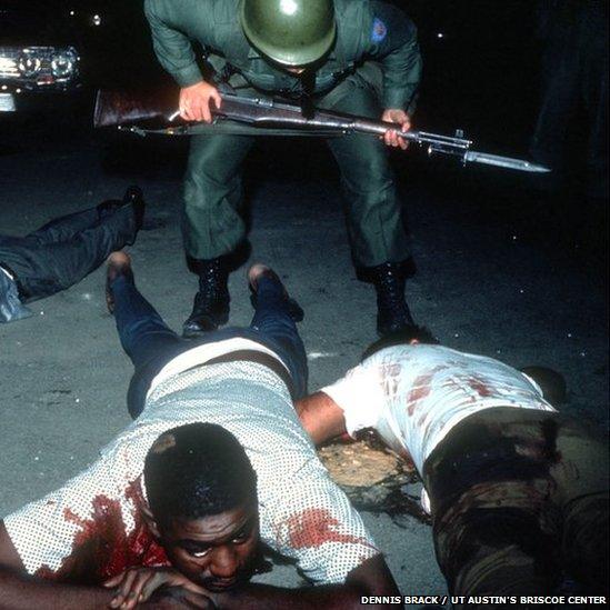 Wounded African American civilians on the ground, bloodied, while a policeman stands above them.