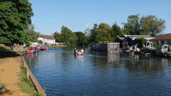 The River Yare flowing through Thorpe St Andrew can be seen, with a boat travelling along it and others moored up