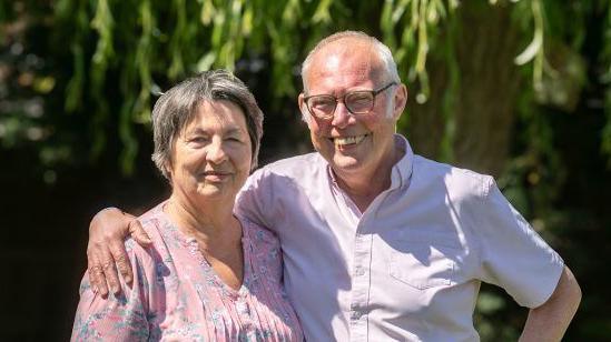 Madge and Ed, standing outside, smiling and looking at the camera. Madge is wearing a floral top, with short hair and Ed, has his arm around Madge, wearing a pink shirt, they are standing in front of a tree.