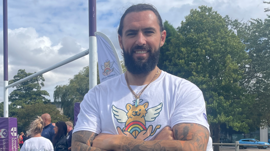 Tom Wilson stood with arms folded. He has tied back dark hair and a beard, and is wearing a white Leo and Friends T-shirt. He is standing in front of a set of rugby goalposts, padded with purple Loughborough University branding.