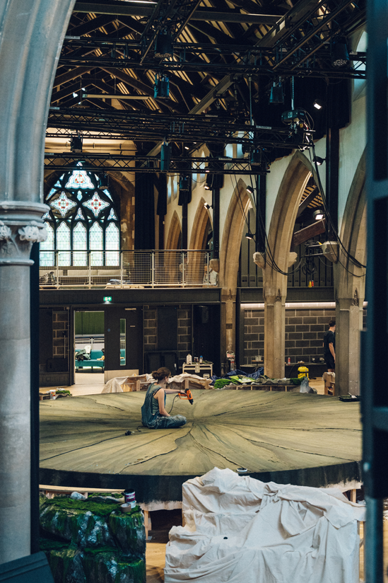 A woman in dungarees drilling into a stone circle one the inside of a church with tall beige arches and a large lighting rig
