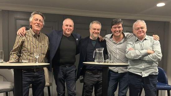 Five men stand in a line, smiling at the camera, in front of two tables with glasses and jugs of water on them, and chairs behind them